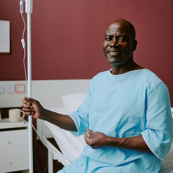 Smiling Elderly African American Man Holding IV Pole Elderly African American man sitting in hospital room, smiling while holding IV pole with one hand, showing positive spirit and comfort in medical setting